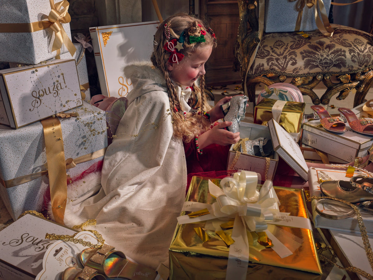 Child wearing a white dress-up cape and festive hair accessories, surrounded by wrapped Christmas gifts.