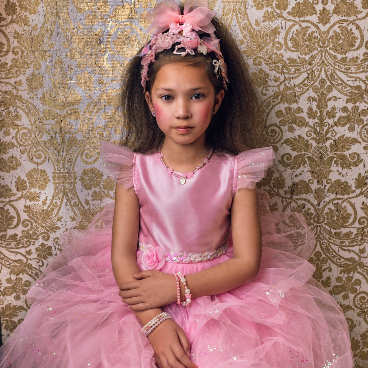A young girl wearing a pink ballgown and matching pink headband sits in front of a gold and beige damask patterned backdrop. The word 'gouza!' is printed at the top.