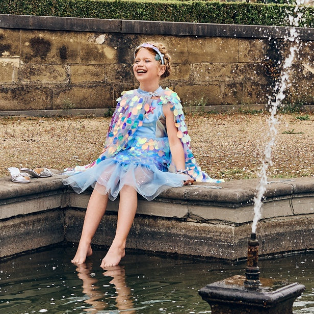 Child in a floral dress sitting by a fountain in an ornate garden.