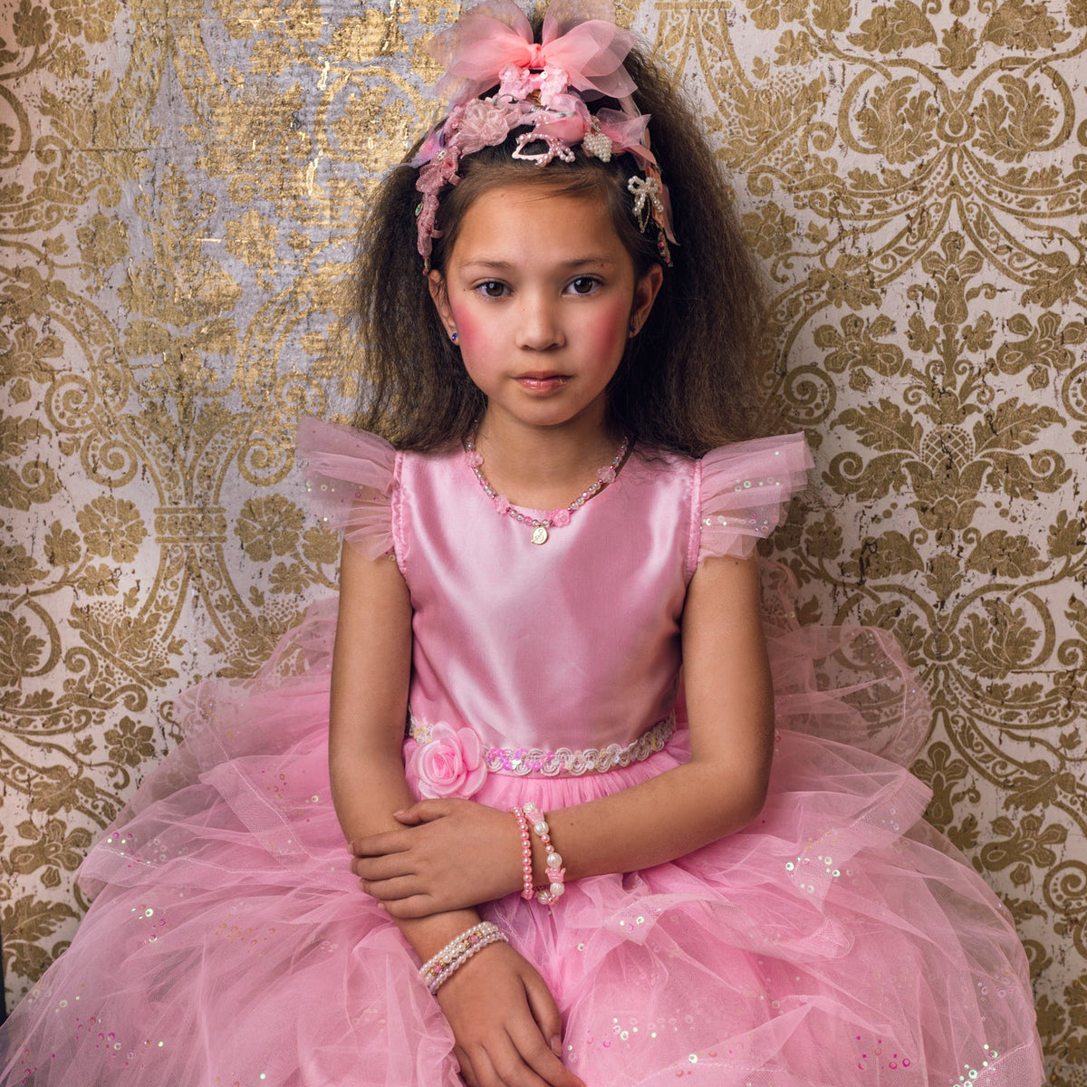 A young girl wearing a pink ballgown and matching pink headband sits in front of a gold and beige damask patterned backdrop. The word 'gouza!' is printed at the top.