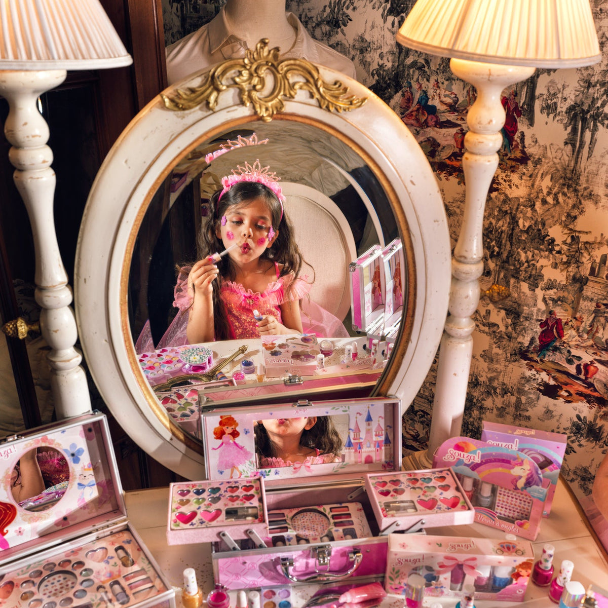 A children's makeup set displayed on a table in front of an ornate mirror, with a young girl visible in the mirror reflection applying makeup. The set includes multiple pink cosmetic palettes, small bottles of nail polish, a gold makeup brush, and other beauty accessories.