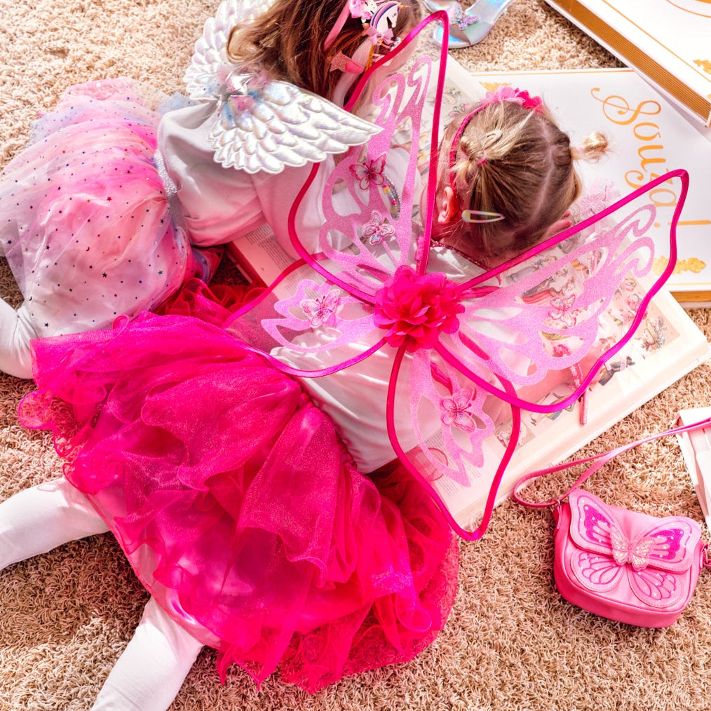 Two young girls lying on a beige carpet, dressed in pink and white fairy costumes with wings. Surrounding them are pink shoes, a small pink purse, a white box with gold trim, and a white booklet.