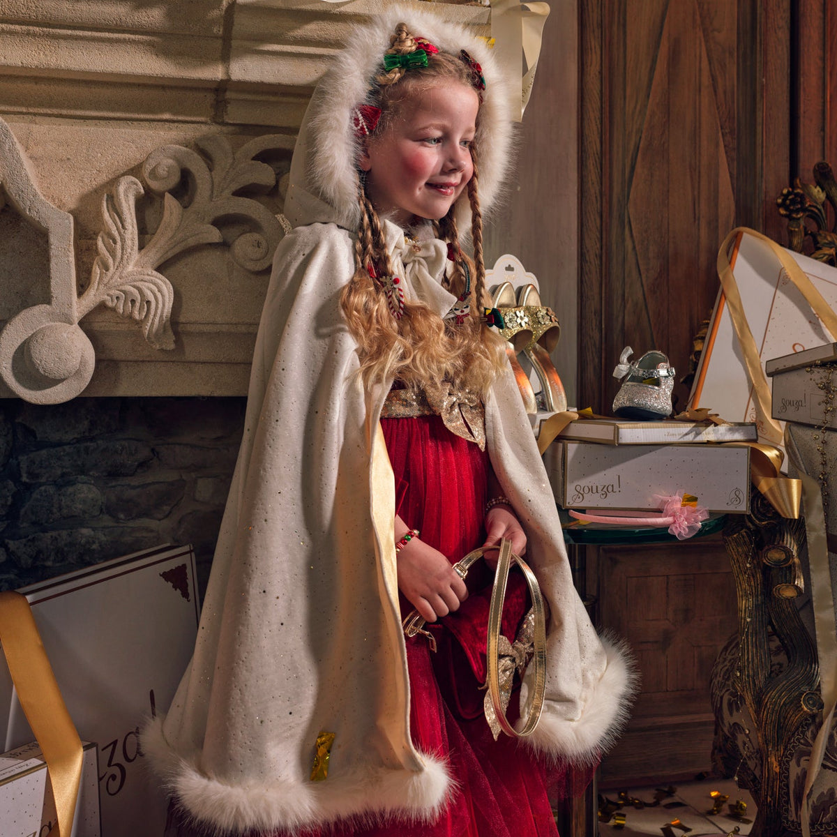 A young girl wearing a white hooded cape with faux fur trim and a red dress, standing in a decorated room with gift boxes and ornaments. The brand name 'gouza!' is visible in gold script in the upper right.
