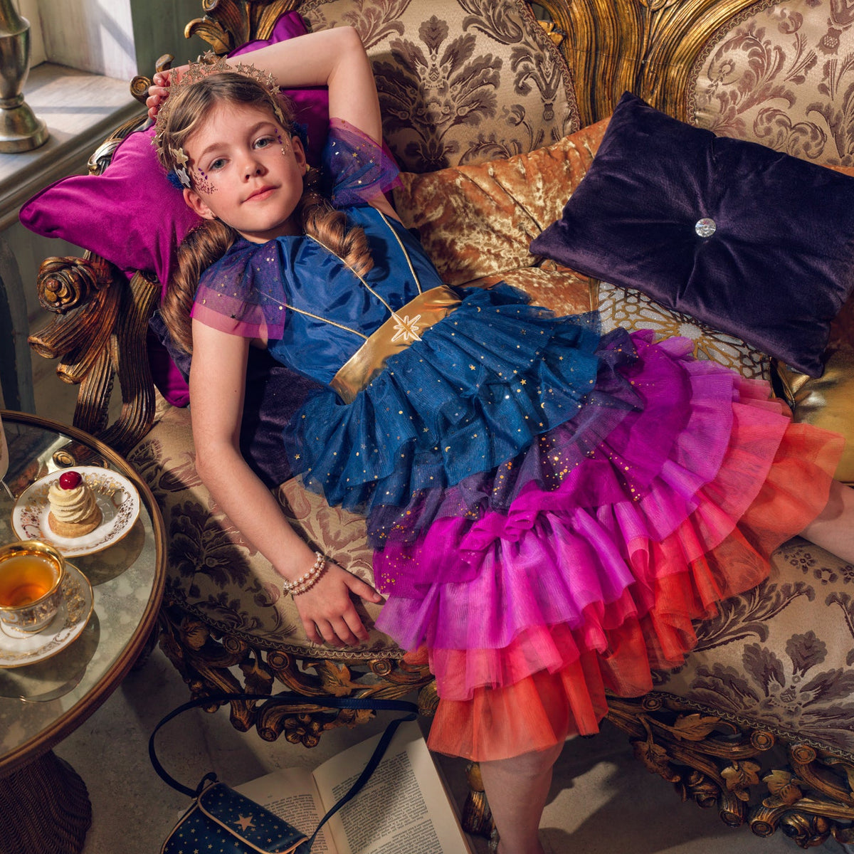 A young girl reclines on an ornate gold and beige sofa wearing a colorful tiered ruffle dress with blue, purple, pink, and orange layers. A small round table beside her holds teacups, a teapot, and pastries in a richly decorated interior.