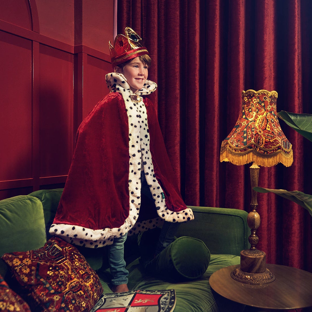 A person wearing a red velvet royal cape with white fur trim and a matching red crown stands on a green sofa in a richly decorated room with red curtains, a patterned table lamp, and green plants.