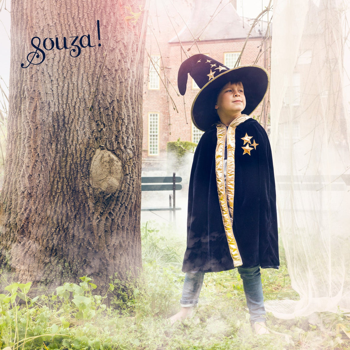 A child wearing a black wizard costume with a gold-trimmed cape, matching pointed hat, and star emblem, standing outdoors in a misty park-like setting next to a large tree trunk. The brand name 'gouza!' appears in the upper left.