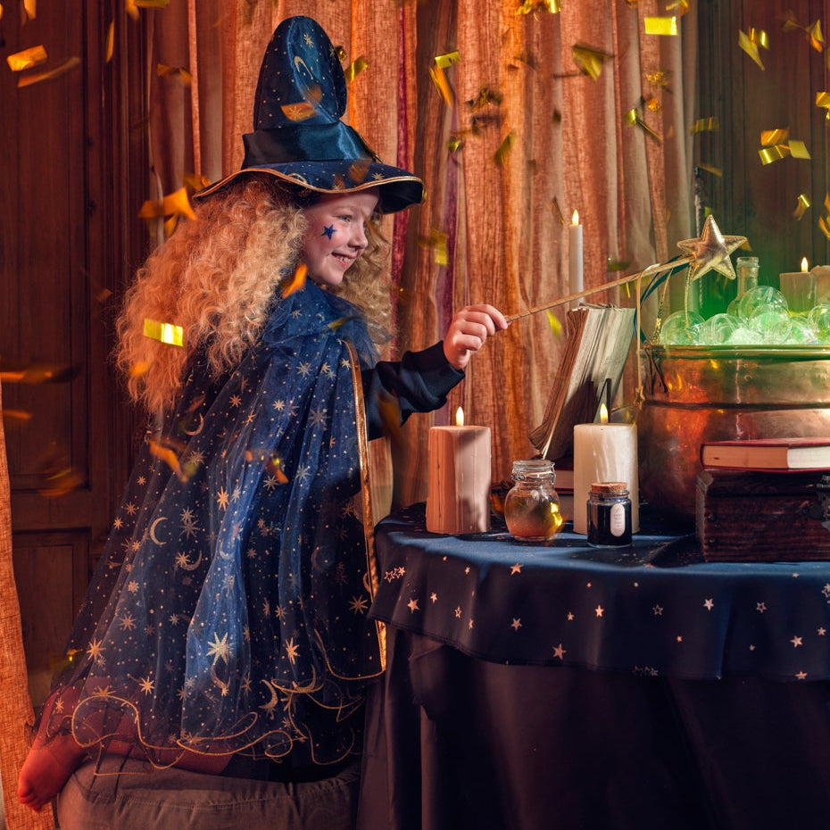 A person dressed as a wizard in a dark blue robe and pointed hat stands beside a table covered with candles and magical props, including a glowing green cauldron, inside a richly decorated room with warm wood paneling, curtains, and falling gold confetti.