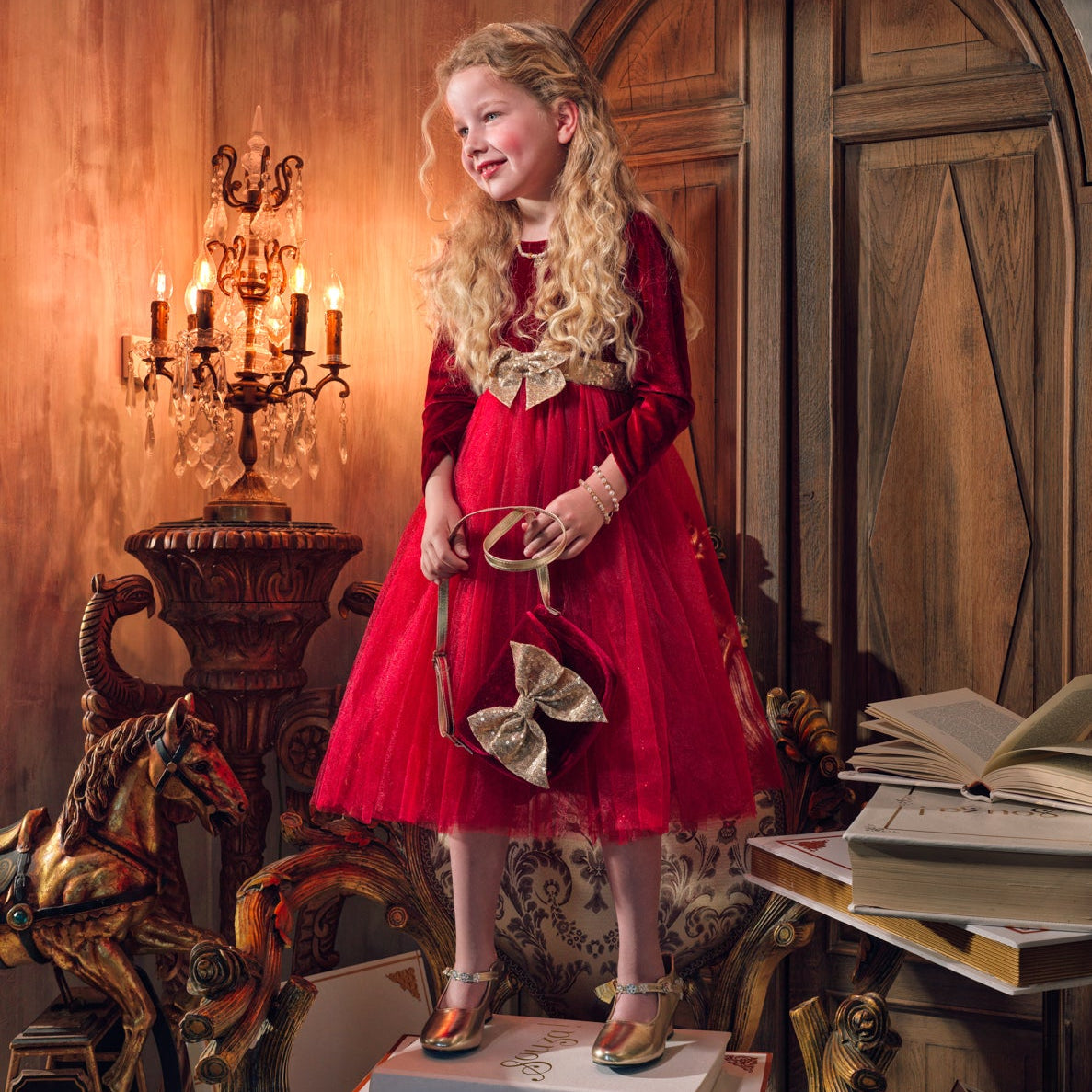 A young girl with long blonde hair stands on books in an ornate vintage room, wearing a red dress with gold trim and holding a gold bow. The room features a large wooden door, a candelabra, and a table with stacked books.