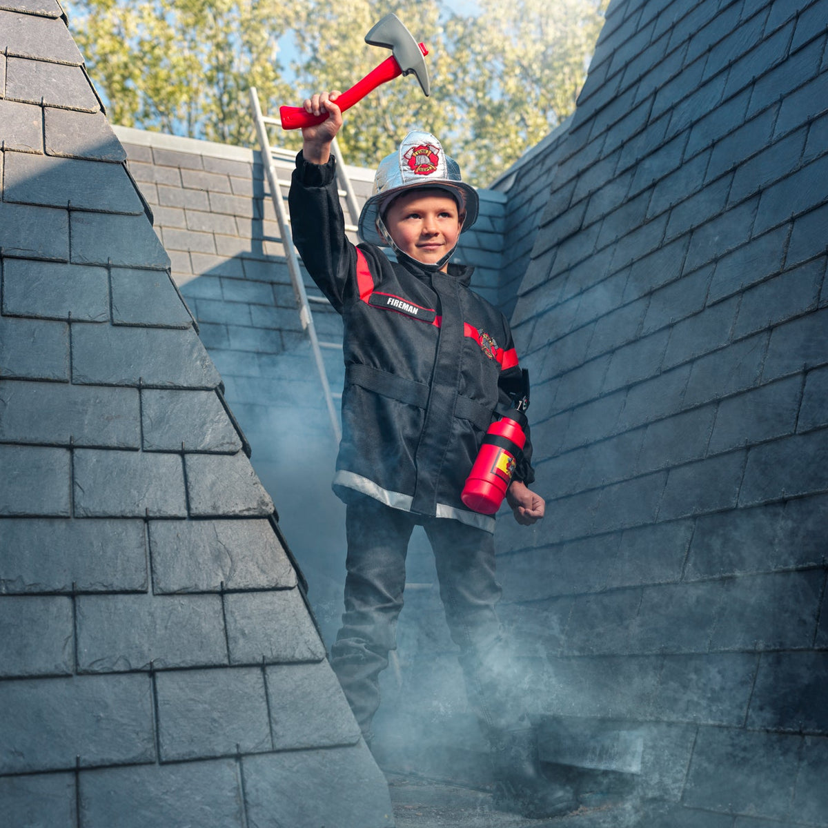 A young boy dressed as a firefighter stands on a gray shingled roof, holding a toy axe and a red fire extinguisher. He is wearing a black firefighter jacket and helmet with reflective details, with smoke effects in the background.