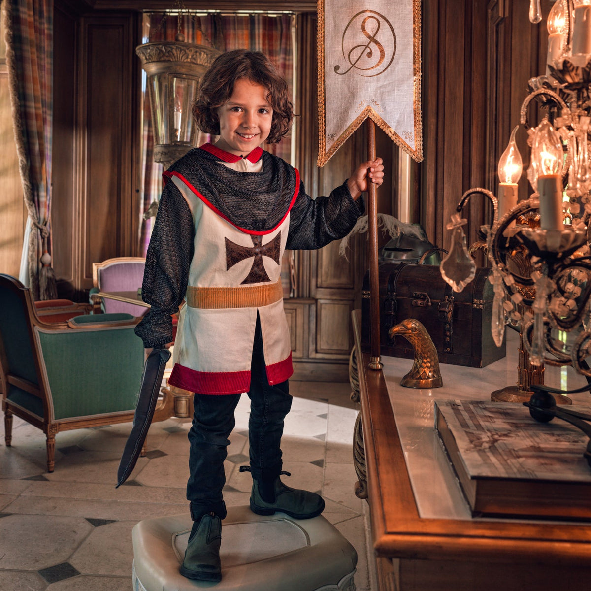 A young boy dressed as a medieval knight stands on a stool in an ornate room, holding a banner with a black cross emblem. He wears a white, red, and black costume with a black chainmail-style top and dark pants.
