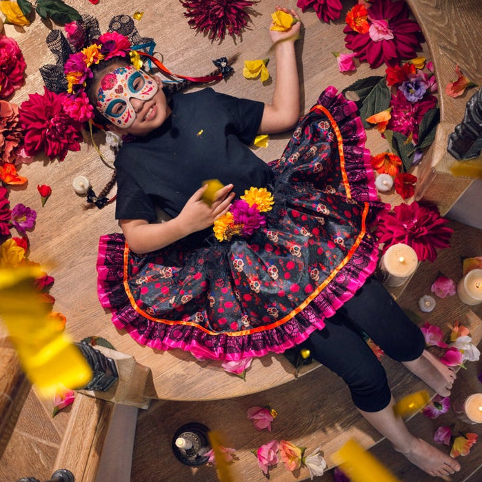 A person wearing a black outfit and a colorful floral skirt lies on a wooden spiral staircase, surrounded by red flowers, candles, petals, and falling yellow confetti.
