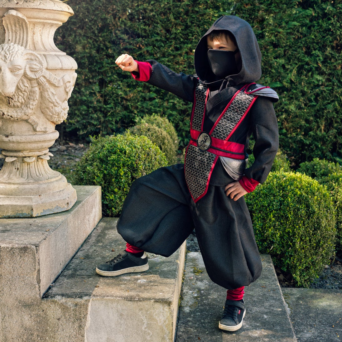 A child wearing a ninja costume with a black hood, face mask, and baggy black pants, standing on stone steps in a garden with trimmed hedges and a large stone planter.