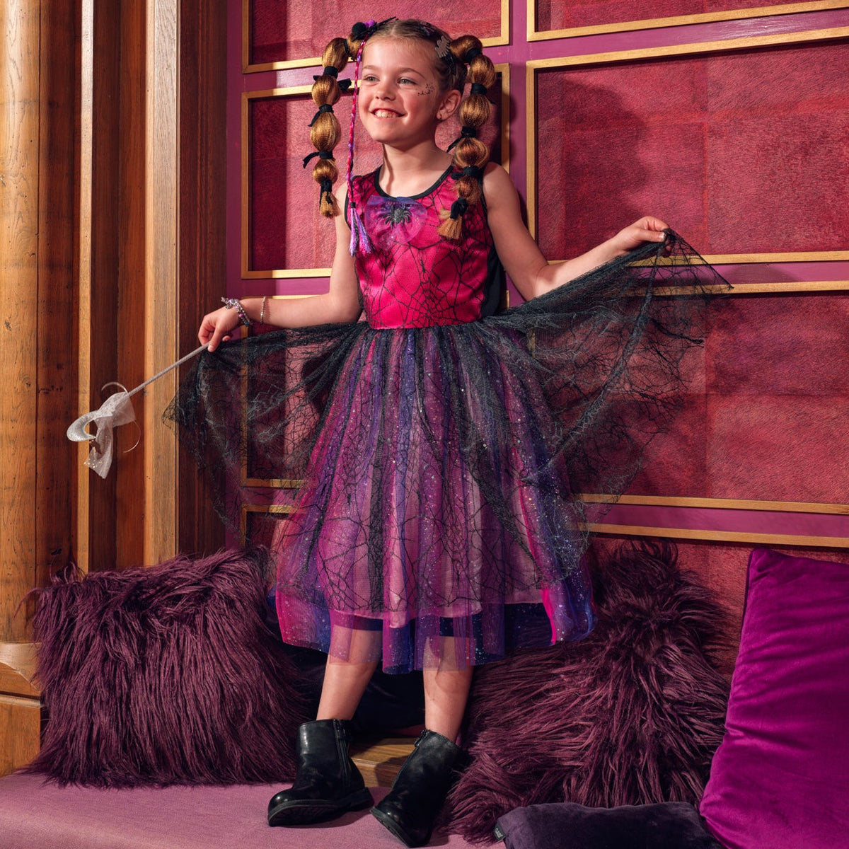 A young girl with curly hair stands on a purple velvet bench in front of a red paneled wall, wearing a sparkly pink and purple tutu dress and holding out a black tulle skirt. She is surrounded by plush purple pillows.