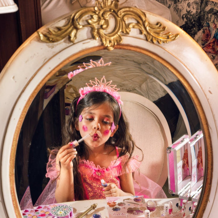 A children's makeup set displayed on a table in front of an ornate mirror, with a young girl visible in the mirror reflection applying makeup. The set includes multiple pink cosmetic palettes, small bottles of nail polish, a gold makeup brush, and other beauty accessories.