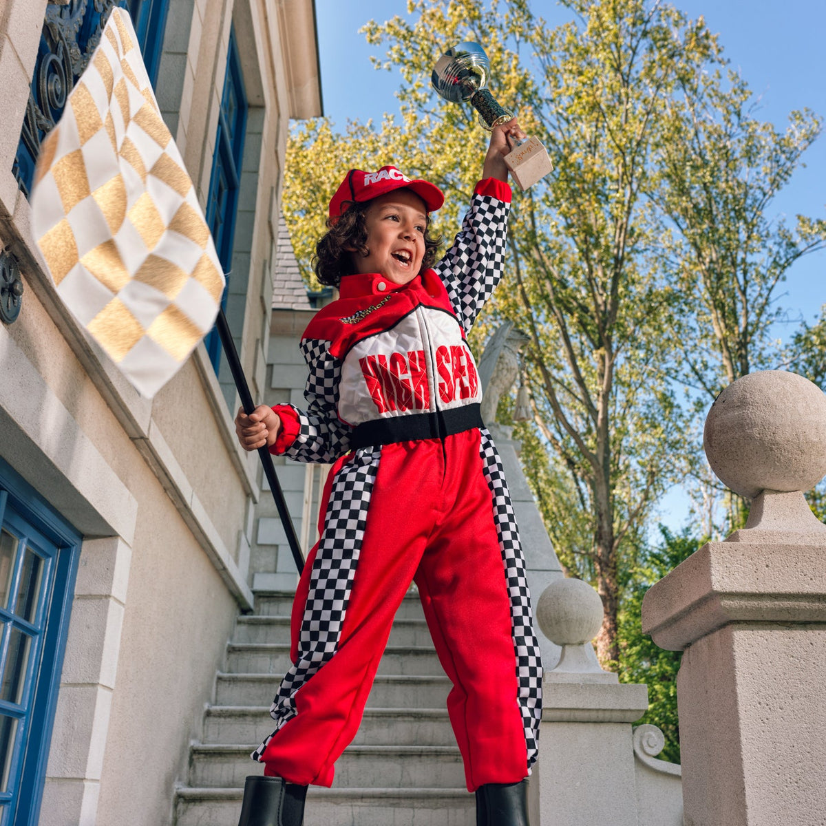 A child wearing a red and black checkered high-speed race costume stands on outdoor stone steps, holding a checkered flag and a trophy. The costume includes a red cap, red jumpsuit with black checkered side panels, a red scarf, and black boots.