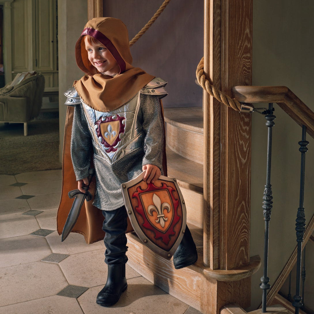 A child wearing a medieval knight costume stands on a staircase landing, holding a shield and sword. The costume includes a gray tunic with a heraldic crest, a brown hood, and a brown cape.