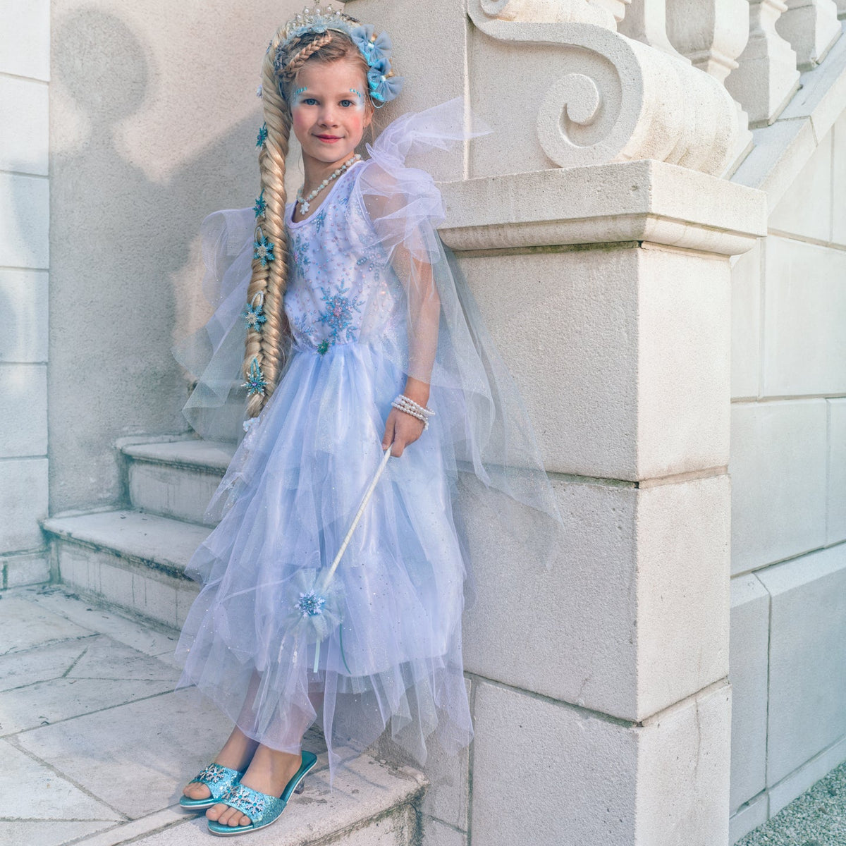 A young girl wearing an Elsa-inspired light blue tulle princess dress with a matching veil, blue floral crown, and long blonde braided wig, standing on stone stairs and holding a wand.